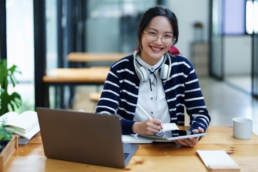 Portrait of a teenage Asian woman using a tablet, wearing headphones and using a notebook to study online via video conferencing on a wooden desk in library.