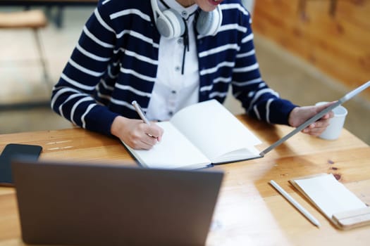 Portrait of a teenage Asian woman using a tablet, wearing headphones and using a notebook to study online via video conferencing on a wooden desk in library.