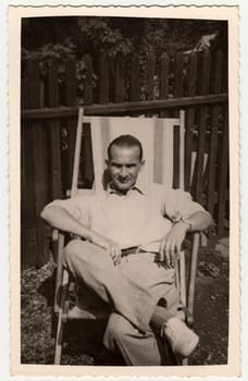 THE CZECHOSLOVAK SOCIALIST REPUBLIC - CIRCA 1950s: Vintage photo shows man sits on wooden/textile deck chair in the garden. Black white photography.