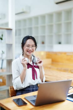 Portrait of a teenage Asian woman using a computer, wearing headphones and using a notebook to study online via video conferencing on a wooden desk in library.