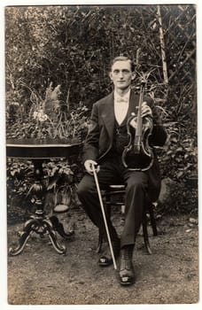 TTHE CZECHOSLOVAK REPUBLIC. - CIRCA 1930s: Vintage photo shows young man poses with the violin outdoors. The violinist sits at the black elegant table. Antique black white photography.