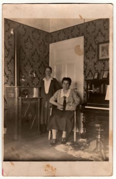 GERMANY - CIRCA 1930s: Vintage photo shows women pose at home. In the picture are piano and stove. Black white antique photography.