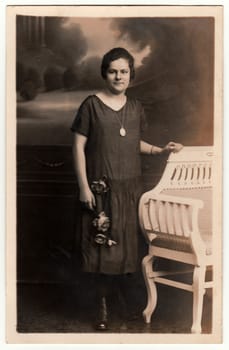 EILENBURG, GERMANY - CIRCA 1930s: Vintage photo shows young woman poses next to historic carved bench. Black white antique studio photography.