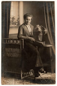 GERMANY - CIRCA 1930s: Vintage photo shows young woman sits on wicker armchair and holds book in the photo studio. Woman sits next to the historic table, on the table is a bouquet. Black white antique studio photography.