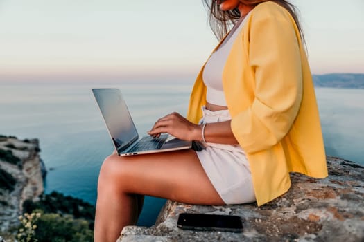 Successful business woman in yellow hat working on laptop by the sea. Pretty lady typing on computer at summer day outdoors. Freelance, travel and holidays concept.