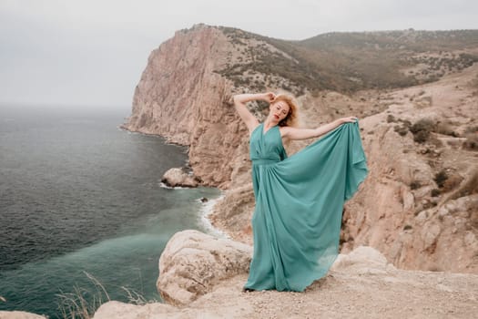 Side view a Young beautiful sensual woman in a mint long dress posing on a volcanic rock high above the sea during sunset. Girl on the nature on overcast sky background. Fashion photo