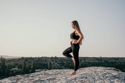 Well looking middle aged woman with long hair, fitness instructor in leggings and tops doing stretching and pilates on the rock near forest. Female fitness yoga routine concept. Healthy lifestyle.