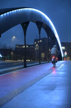 Woman with gray jacket riding a bicycle in rain over illuminated highway bridge in Heerdt