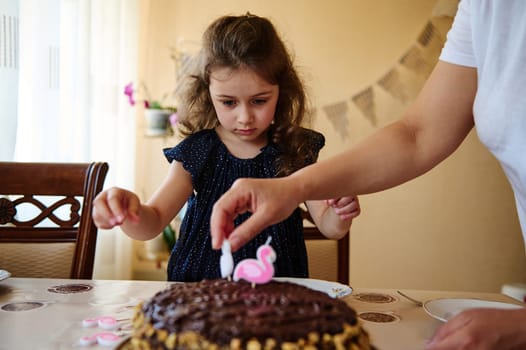 Adorable 5 years old Caucasian child, lovely birthday girl in elegant navy dress with golden dots, sitting at a dining table and looking at her mom's hand putting candles on birthday cake.