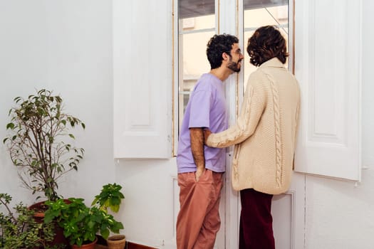 happy couple of gay men looking out the window of their home, concept of real people lifestyle and love between people of the same sex