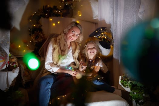 Cute mother and daughter in a room with Christmas garlands. The tradition of decorating the house and dressing up for the holidays. Gifts for family. Happy childhood and motherhood