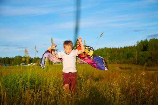 Handsome boy with bright butterfly wings having fun in meadow on natural landscape with grass and flowers on sunny summer day. Portrait of teenage guy in spring season outdoors on field