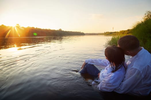 Beautiful adult couple having hugs and fun on nature in the water of a river or lake in the summer evening at sunset. A guy and a girl swim and relax outdoors in clothes in white shirts and jeans