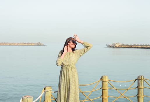 A young beautiful woman in a long dress stands alone on a pier by the sea, looking at the sun