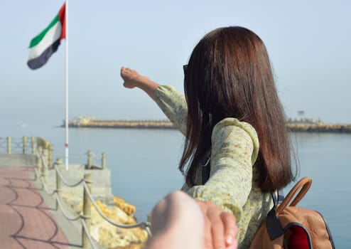 A young woman tourist in a long dress with a backpack points with her hand the direction to the sea to the flag of the United Arab Emirates, and leads her to the pier