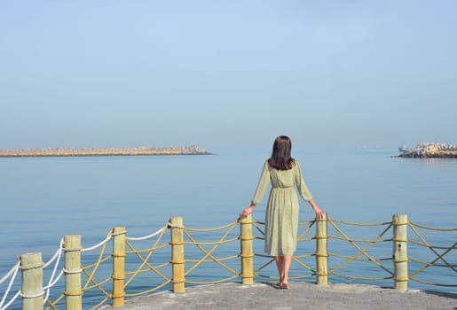 A young woman in a long dress with a backpack walks along the pier by the sea against the background of the developing UAE flag, and looks into the distance