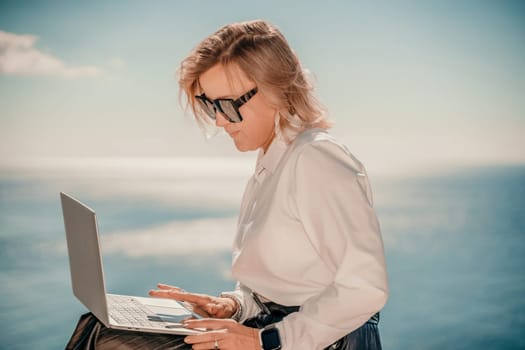 Business woman on nature in white shirt and black skirt. She works with an iPad in the open air with a beautiful view of the sea. The concept of remote work