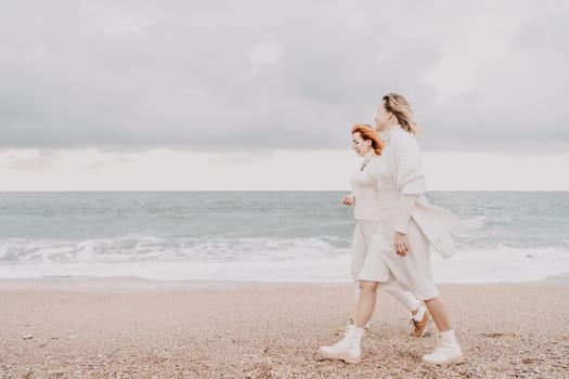 Women sea walk friendship spring. Two girlfriends, redhead and blonde, middle-aged walk along the sandy beach of the sea, dressed in white clothes. Against the backdrop of a cloudy sky and the winter sea. Weekend concept