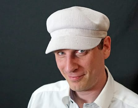 Portrait of a man with a white baker boy cap looking into the camera. Dark grey background.