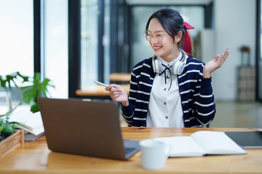 Portrait of a teenage Asian woman using a computer, wearing headphones and using a notebook to study online via video conferencing on a wooden desk in library.