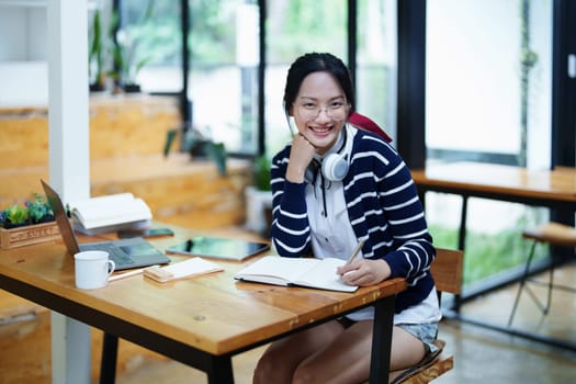 Portrait of a teenage Asian woman using a computer, wearing headphones and using a notebook to study online via video conferencing on a wooden desk in library.