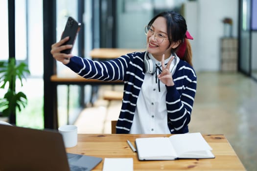 Portrait of a teenage Asian woman using a smartphone, wearing headphones and using a notebook to study online via video conferencing on a wooden desk in library.