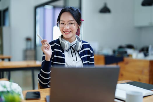 Portrait of a teenage Asian woman using a computer, wearing headphones and using a notebook to study online via video conferencing on a wooden desk in library.