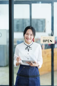 Starting and opening a small business, a young Asian woman showing a smiling face holding a tablet in an apron standing in front of a coffee shop bar counter. Business Owner, Restaurant, Cafe concept.