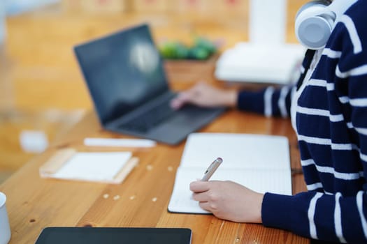 Portrait of a teenage Asian woman using a computer, wearing headphones and using a notebook to study online via video conferencing on a wooden desk in library.