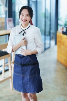 Starting and opening a small business, a young Asian woman showing a smiling face holding a tablet in an apron standing in front of a coffee shop bar counter. Business Owner, Restaurant, Cafe concept.