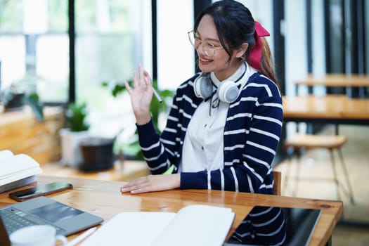 Portrait of a teenage Asian woman using a computer, wearing headphones and using a notebook to study online via video conferencing on a wooden desk in library.