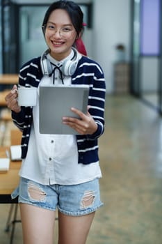 Portrait of a teenage Asian woman using a tablet, wearing headphones and using a notebook to study online via video conferencing on a wooden desk in library.