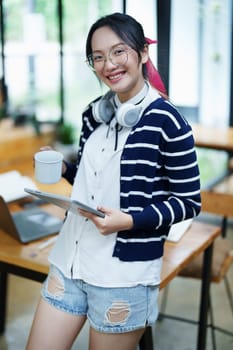 Portrait of a teenage Asian woman using a tablet, wearing headphones and using a notebook to study online via video conferencing on a wooden desk in library.