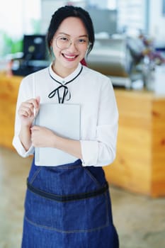 Starting and opening a small business, a young Asian woman showing a smiling face holding a tablet in an apron standing in front of a coffee shop bar counter. Business Owner, Restaurant, Cafe concept.