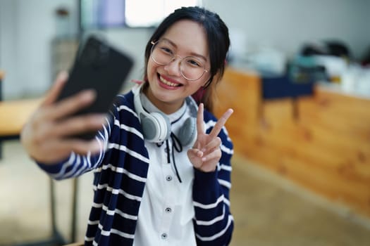 Portrait of a teenage Asian woman using a smartphone, wearing headphones and using a notebook to study online via video conferencing on a wooden desk in library.