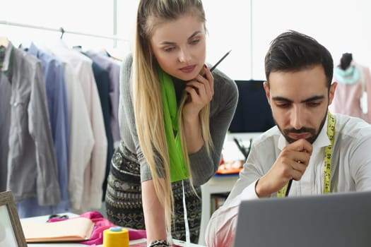 Two fashion designers work as a team at table and look thoughtfully at computer monitor. Teamwork in creative fashion atelier workshop