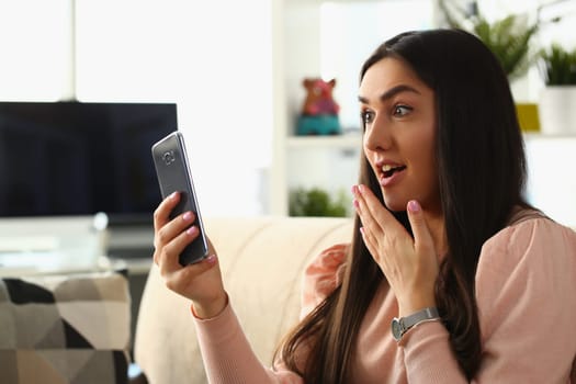 Woman surprised in shock looks at smartphone screen while sitting on couch at home. Emotion joy delight and good news concept