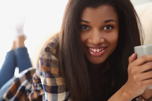 Young beautiful afro woman drinks coffee and lies on sofa. Home leisure and joyful mood concept