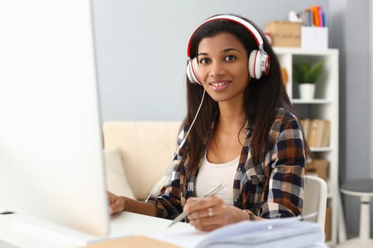 African female student wears headphones studying alone at home office desk. Mixed race woman listening to audio podcast and learning english language concept and taking notes concept