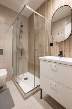 Diagonal shot of small bathroom with stylish white vanity glass shower and ceramic sink with beautiful beige tiles. Concept bathroom after renovation in the apartment.