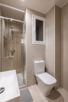 Multifunctional small bathroom with stylish beige tiles, glass shower door, washbasin, toilet and window. Concept of modern bathroom in a hotel. Single persons apartment.