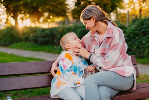 mother and son sit on a park bench in the rays of the setting sun. the concept of a family. Mother's Day. beautiful girl (mother) with a boy (son) in the park in the park are sitting on a bench at sunset.