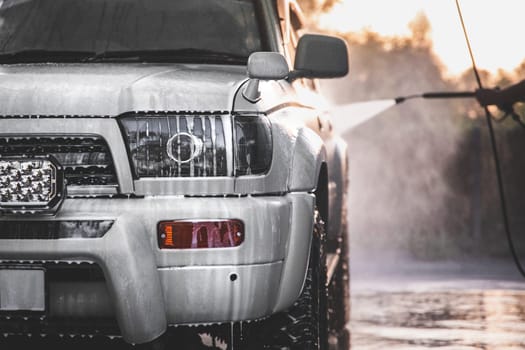 Washing white car at a self-service car wash with a high-pressure water jet after off-road driving.