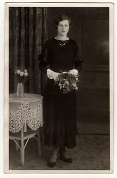 BERLIN, GERMANY - CIRCA 1940s: Vintage photo shows woman wears black dress and she holds bouquet in hands. Studio photography contains the small table and crocheted tablecloth.