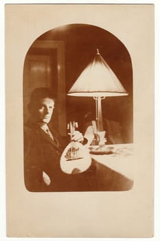 CHEMNITZ, GERMANY - MAY 10, 1927: Vintage photo shows man with the mandolin. He sits at the table. The period lamp is on the background.