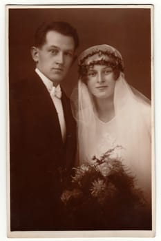 THE CZECHOSLOVAK REPUBLIC - CIRCA 1930s: Vintage photo of newlyweds. Bride wears a long veil and holds wedding bouquet. Groom wears black suit and white bow tie. Black white antique studio portrait.