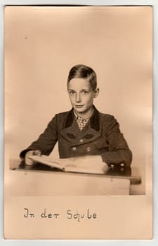 GERMANY - AUGUST 6, 1942: Vintage photo shows young boy (pupil) at the school desk. Pupil wears a period jacket (oaken epaulettes/marks on jacket). On photography is text in German - "In the school" (translation in English).