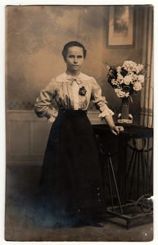 THE CZECHOSLOVAK REPUBLIC - CIRCA 1920s: Vintage photo shows woman wears an elegant dress, poses next to black table. On the table is vase with flowers. Black & white antique studio photography with sepia effect.