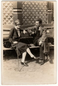 LUHACOVICE, THE CZECHIOSLOVAK REPUBLIC - CIRCA 1930s: Vintage photo shows an elegant couple sits on the bench at spa resort. Black & white antique photography.