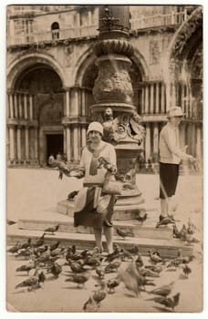 VENEZIA, ITALY - 1926: Vintage photo shows an elegant woman holds pigeons in hands. Second woman feeds pigeons/doves with grain. Women on holiday (vacation).